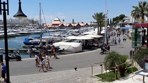 A marina with boats, a covered area with tables and umbrellas, and a pedestrian walkway with people and palm trees. - Aldeia do Golf- Family with Pool and Wi-fi (Vilamoura)