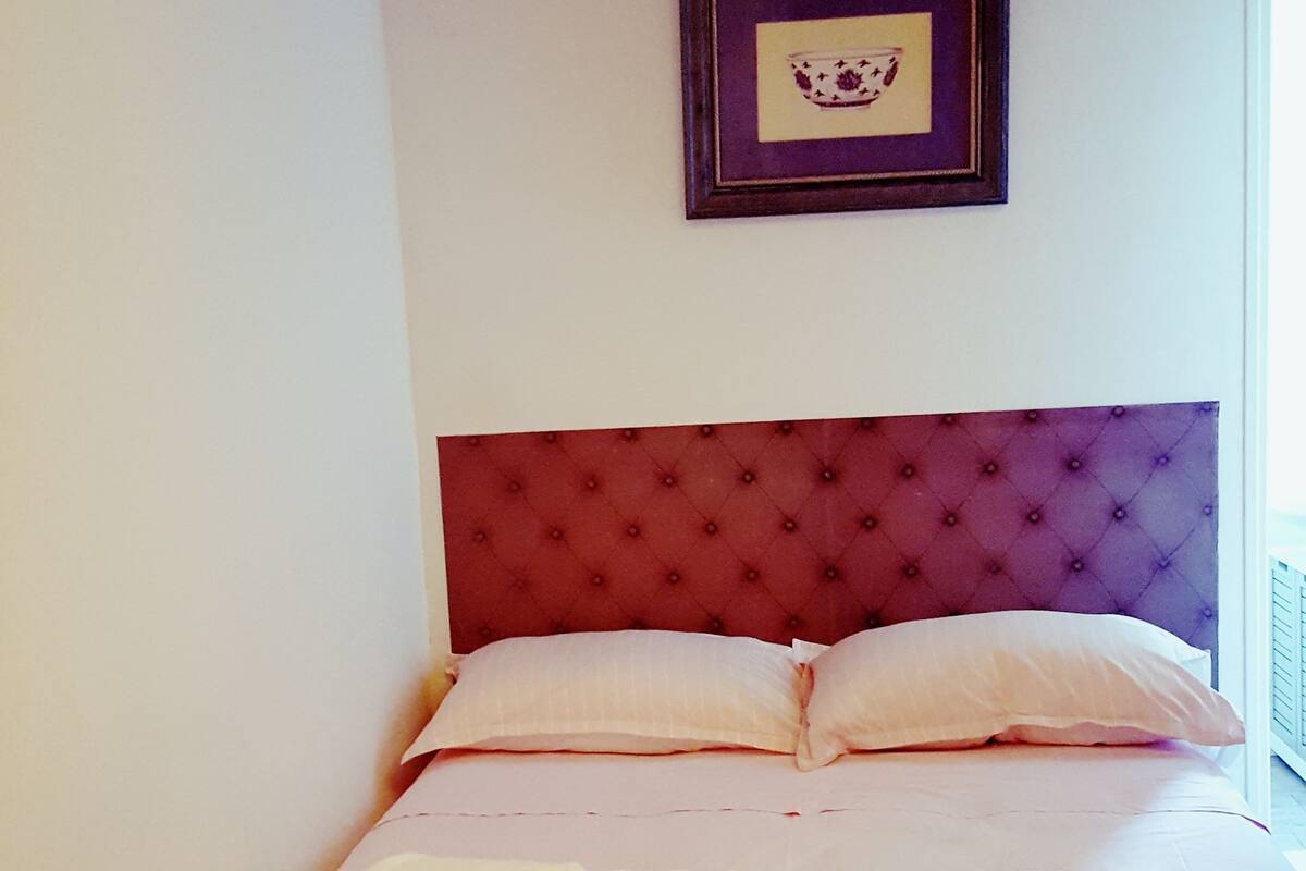 A bed with a tufted headboard, a framed picture above it, and a window to the right.