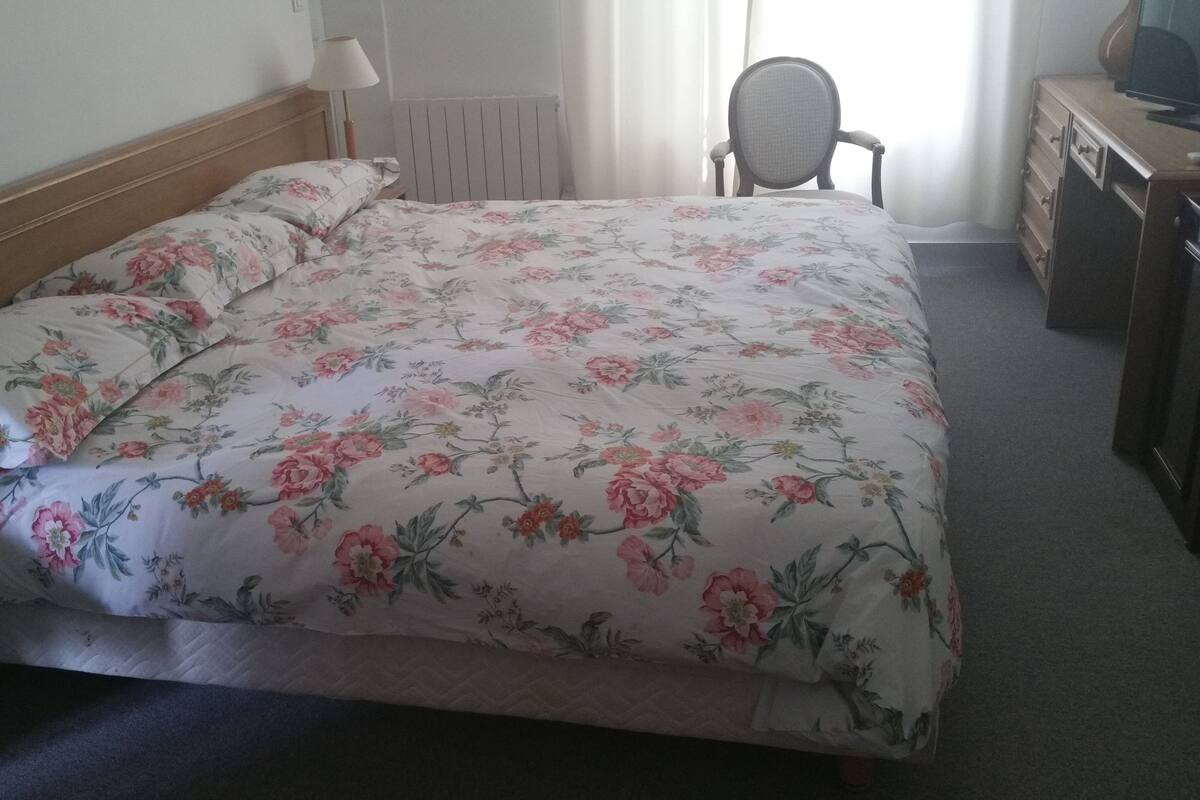 A neatly made bed with floral bedding, a wooden headboard, a bedside table with a lamp, and a chair in the corner.
