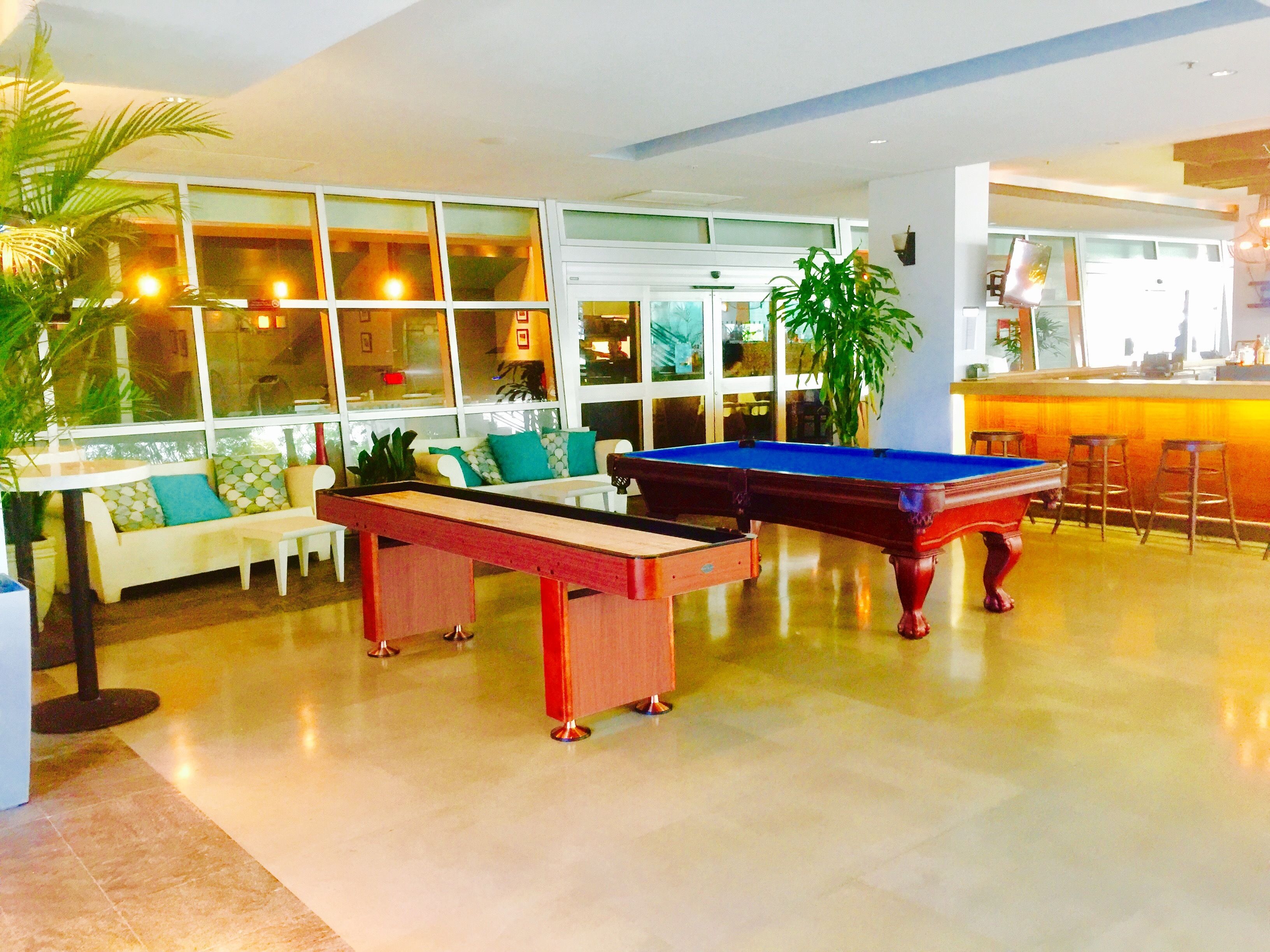 A pool table in a well-lit indoor space with large windows, a sofa with patterned cushions, and a bar area.