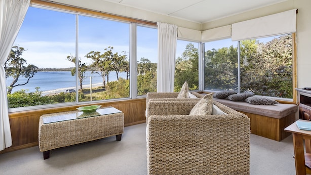 A room with large windows offering a view of the sea, wicker furniture, and a glass-top coffee table. - Summertime Beach Cottage (Southport)