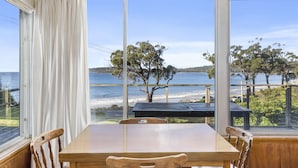 A dining area with a wooden table and chairs, large windows with a view of the beach and ocean. - Summertime Beach Cottage (Southport)