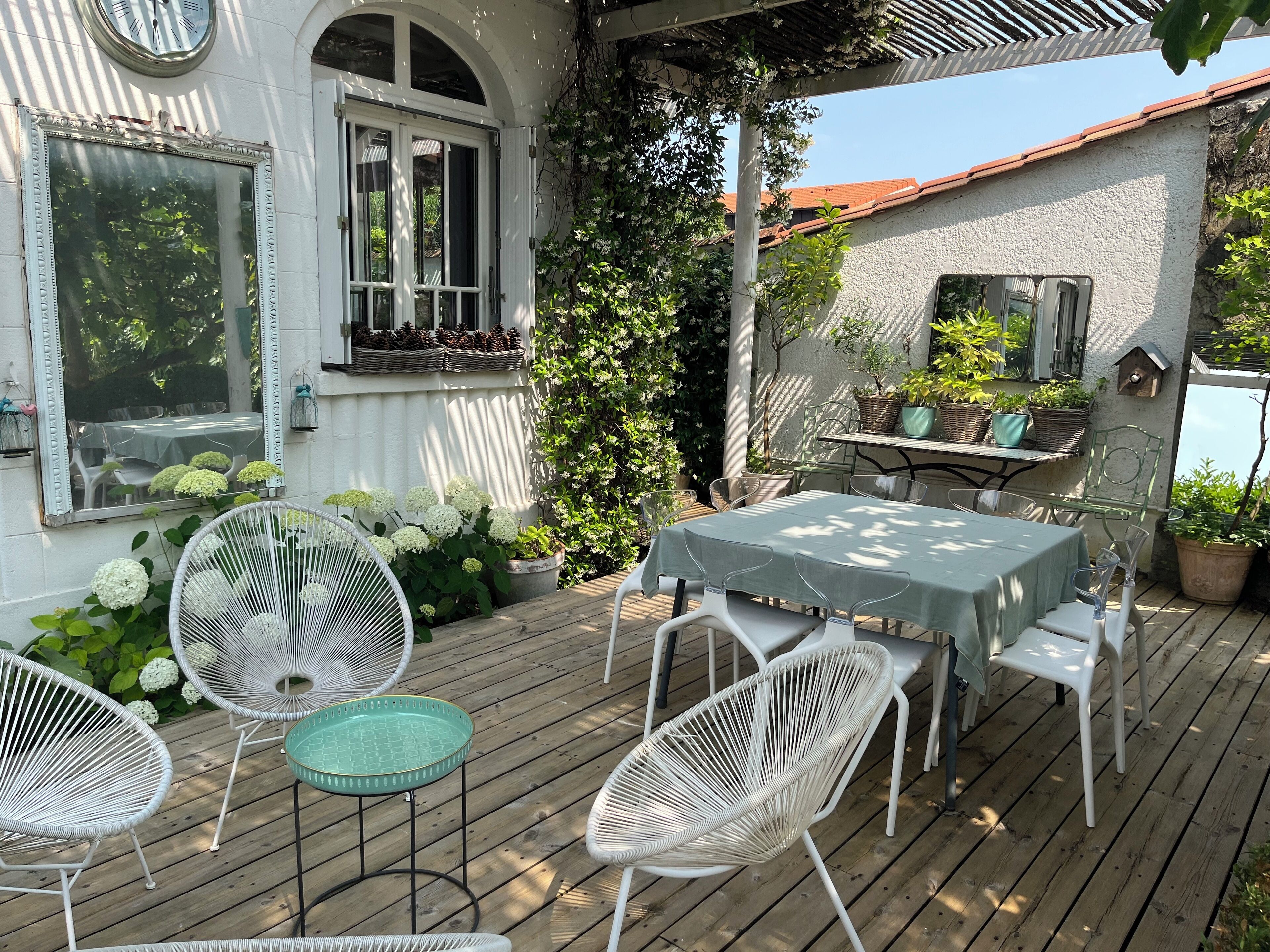 Una terraza de madera con una mesa y sillas, rodeada de plantas en macetas y un edificio blanco con una ventana y una puerta.
