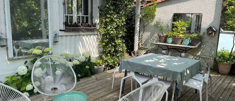 Una terraza de madera con una mesa y sillas, rodeada de plantas en macetas y un edificio blanco con una ventana y una puerta.