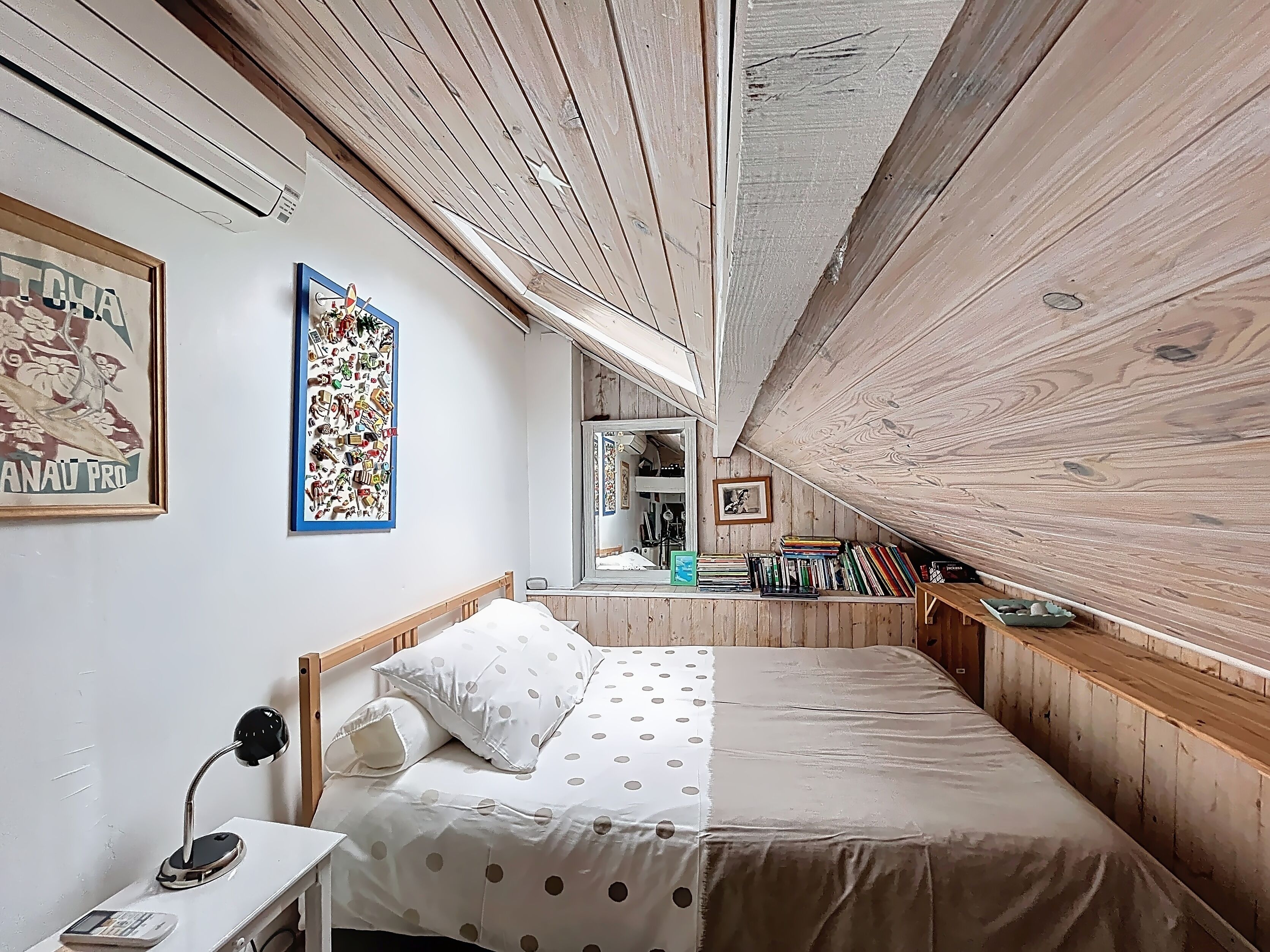 A bedroom with a wooden ceiling, a bed with polka dot bedding, a desk with a lamp, and bookshelves filled with books.
