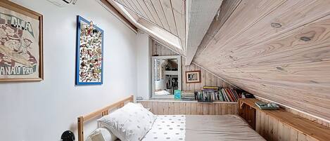 A bedroom with a wooden ceiling, a bed with polka dot bedding, a desk with a lamp, and bookshelves filled with books.