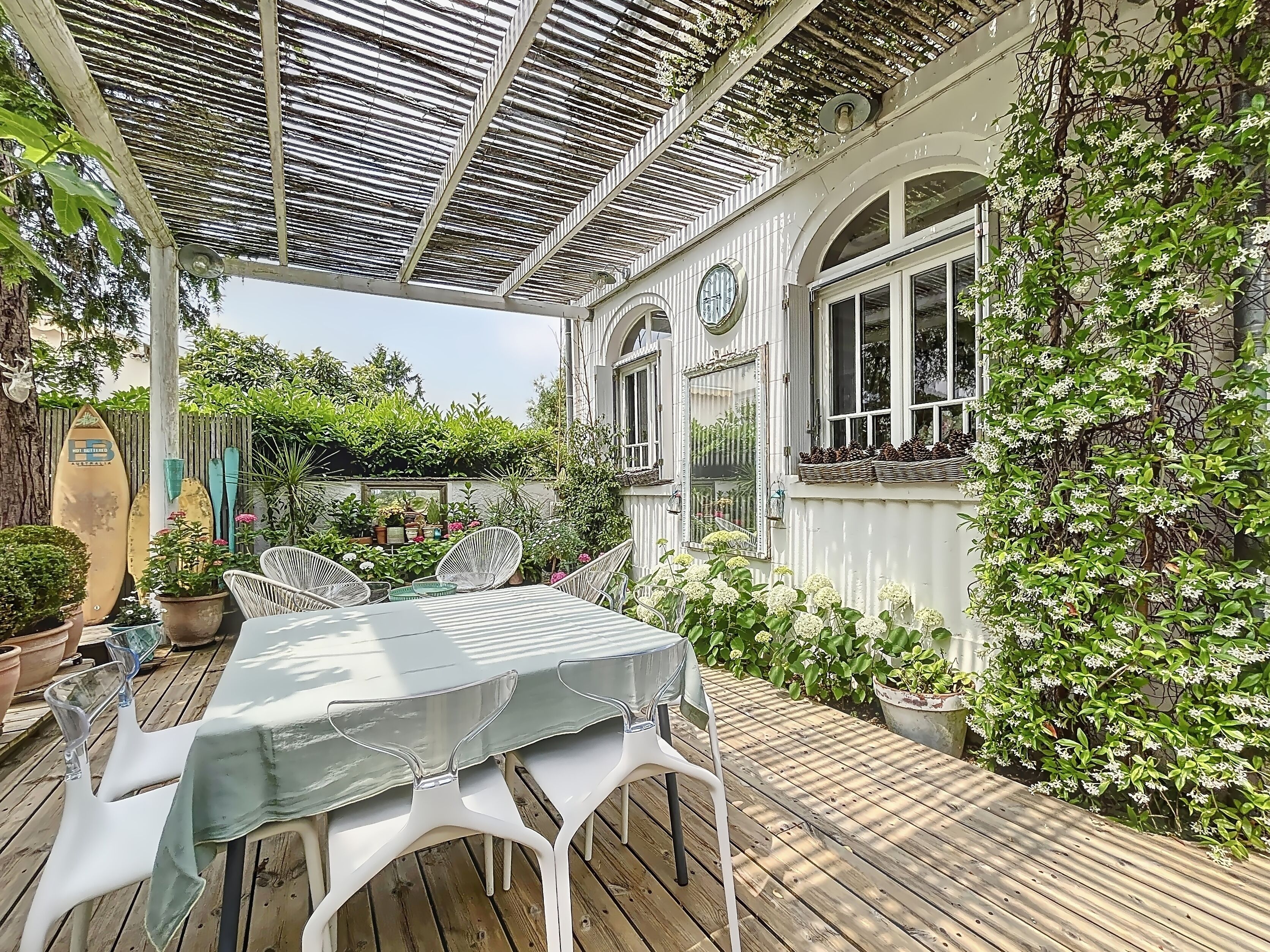 Une terrasse couverte avec une table et des chaises, entourée d’une végétation luxuriante et de plantes en pot.
