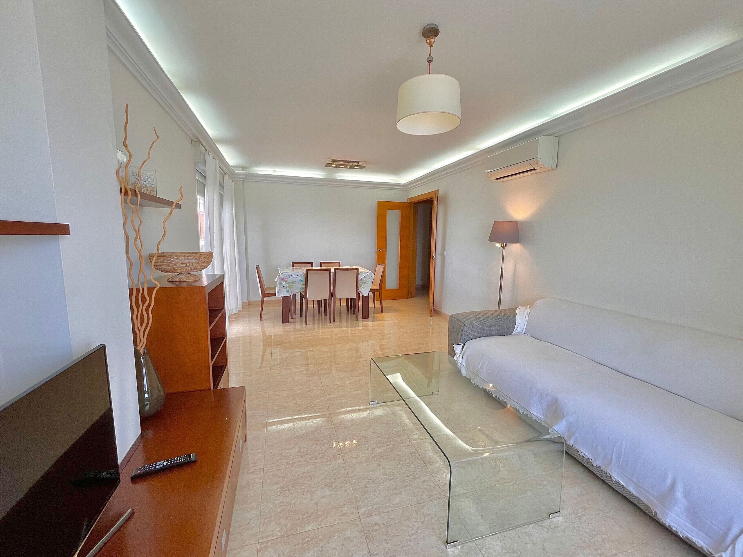 Una sala de estar moderna con una mesa de centro de vidrio, un sofá, un comedor con una mesa y sillas, y una lámpara.