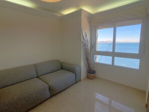 Ein Wohnzimmer mit einem beigefarbenen Sofa, einer Topfpflanze und einem großen Fenster mit Meerblick.