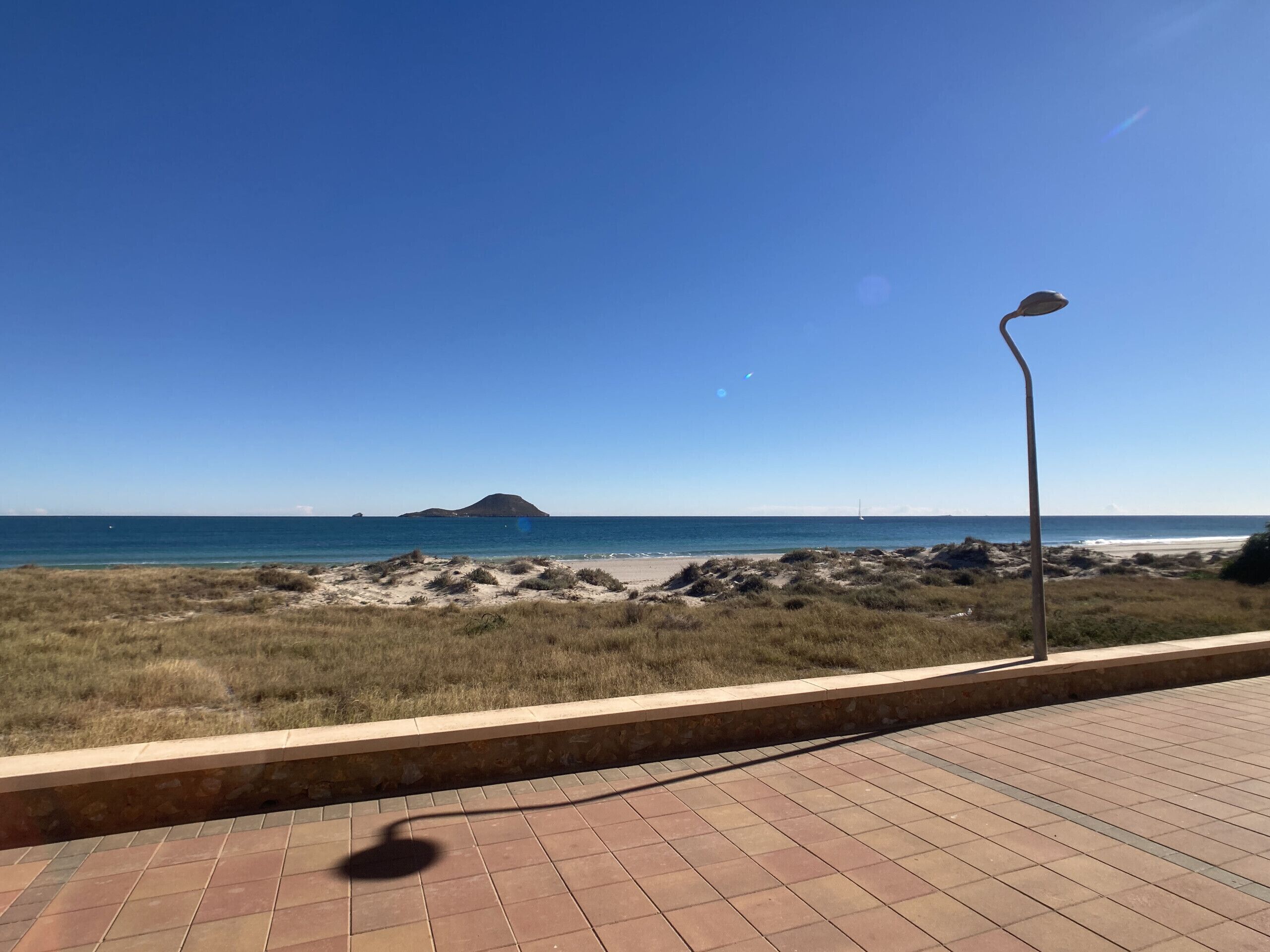Un camino pavimentado con una farola, que conduce a una playa con costa rocosa y una isla en la distancia.