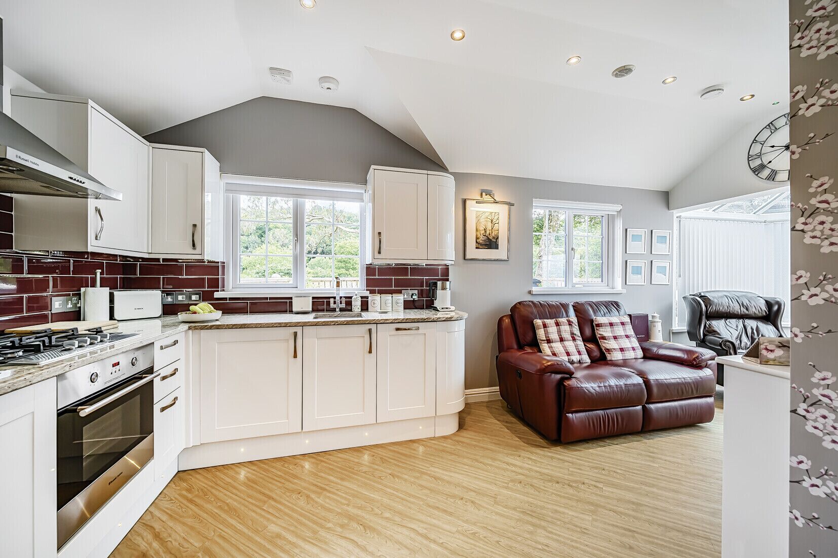 A modern kitchen with white cabinets, a built-in oven, and a gas stove. There is a brown leather sofa with cushions and a black leather chair.