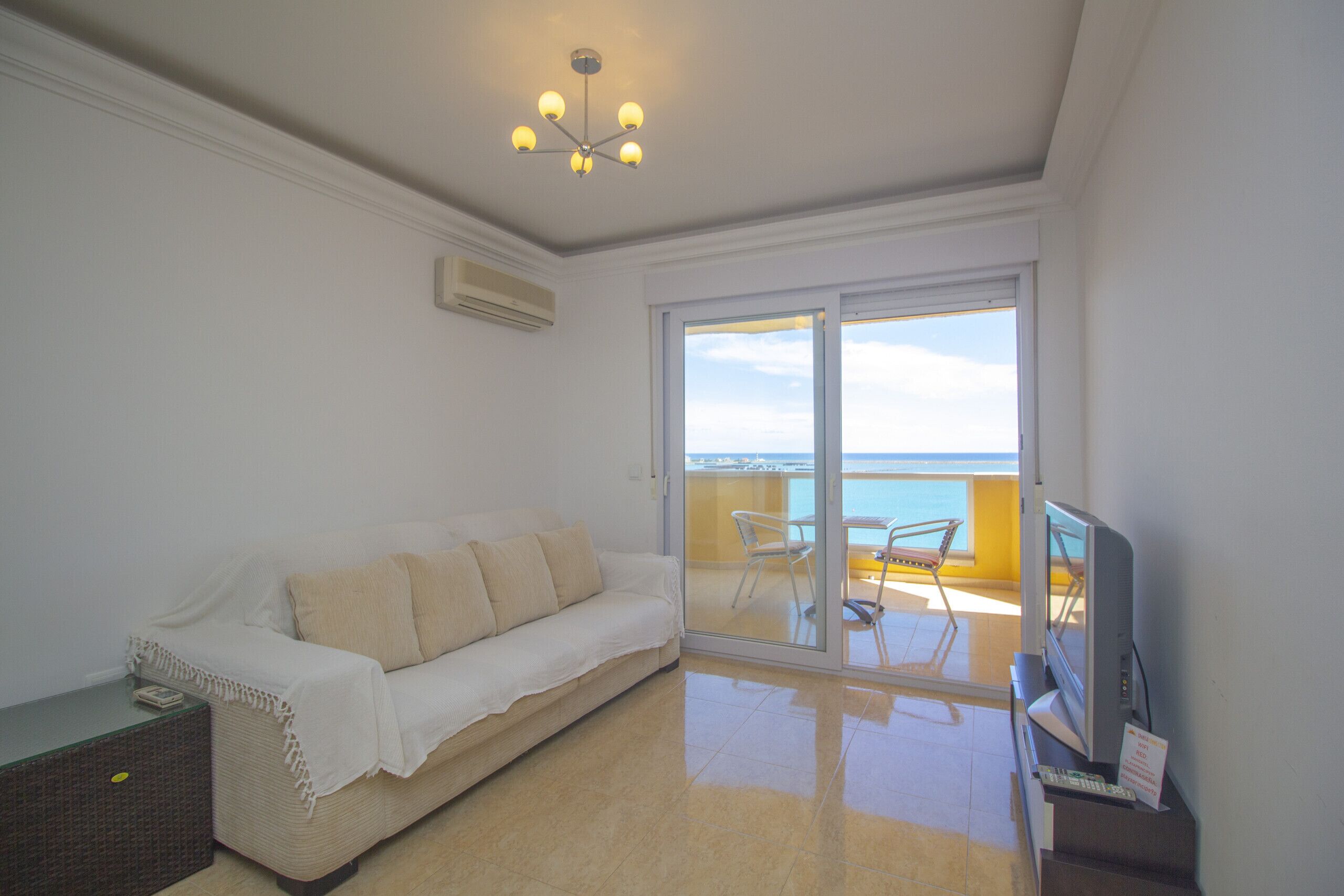 A living room with a sofa, a television, and a balcony with a view of the sea.