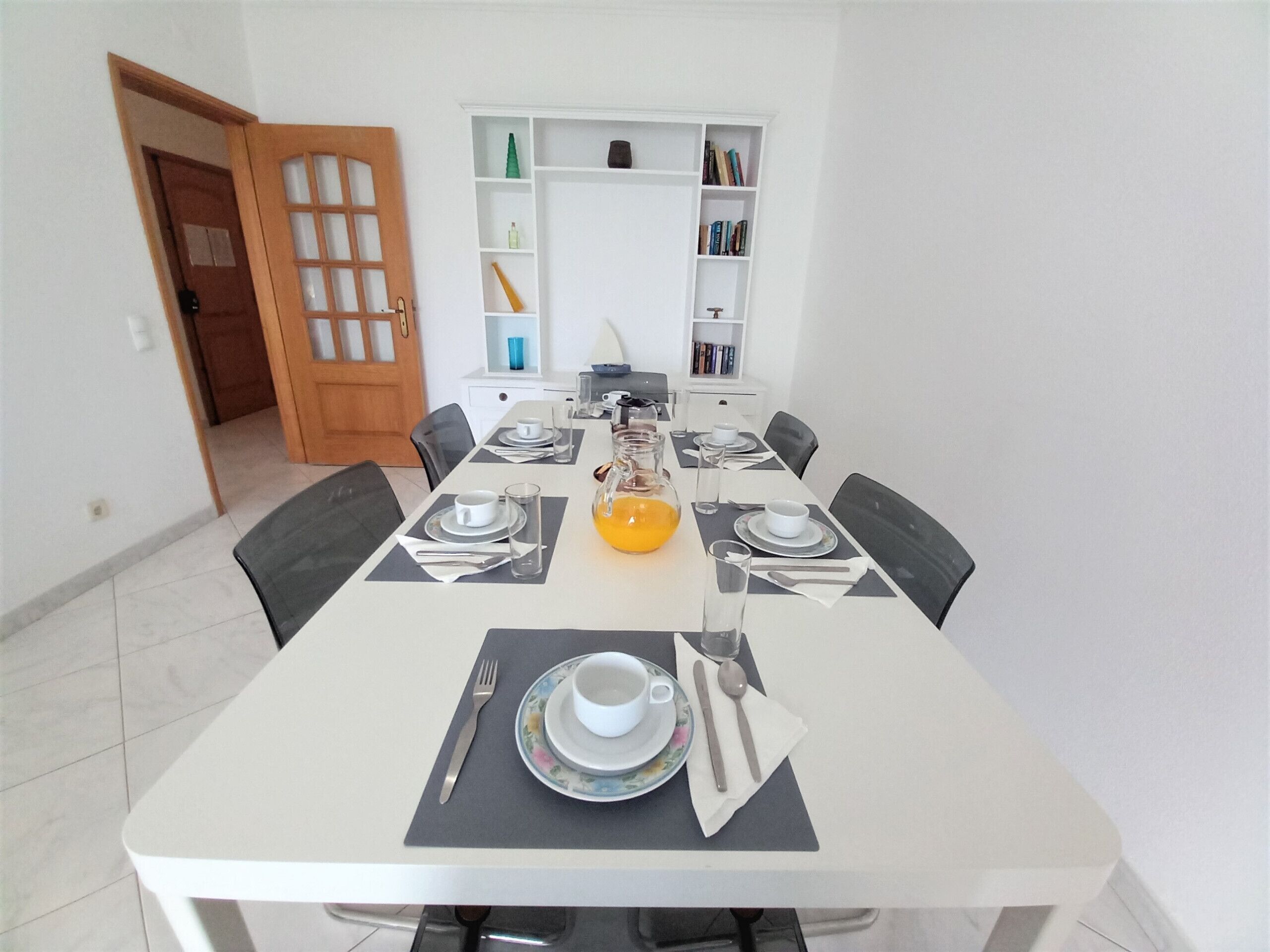 Un coin repas avec une table blanche dressée pour quatre personnes, comprenant des assiettes, des couverts et une carafe de jus d’orange.