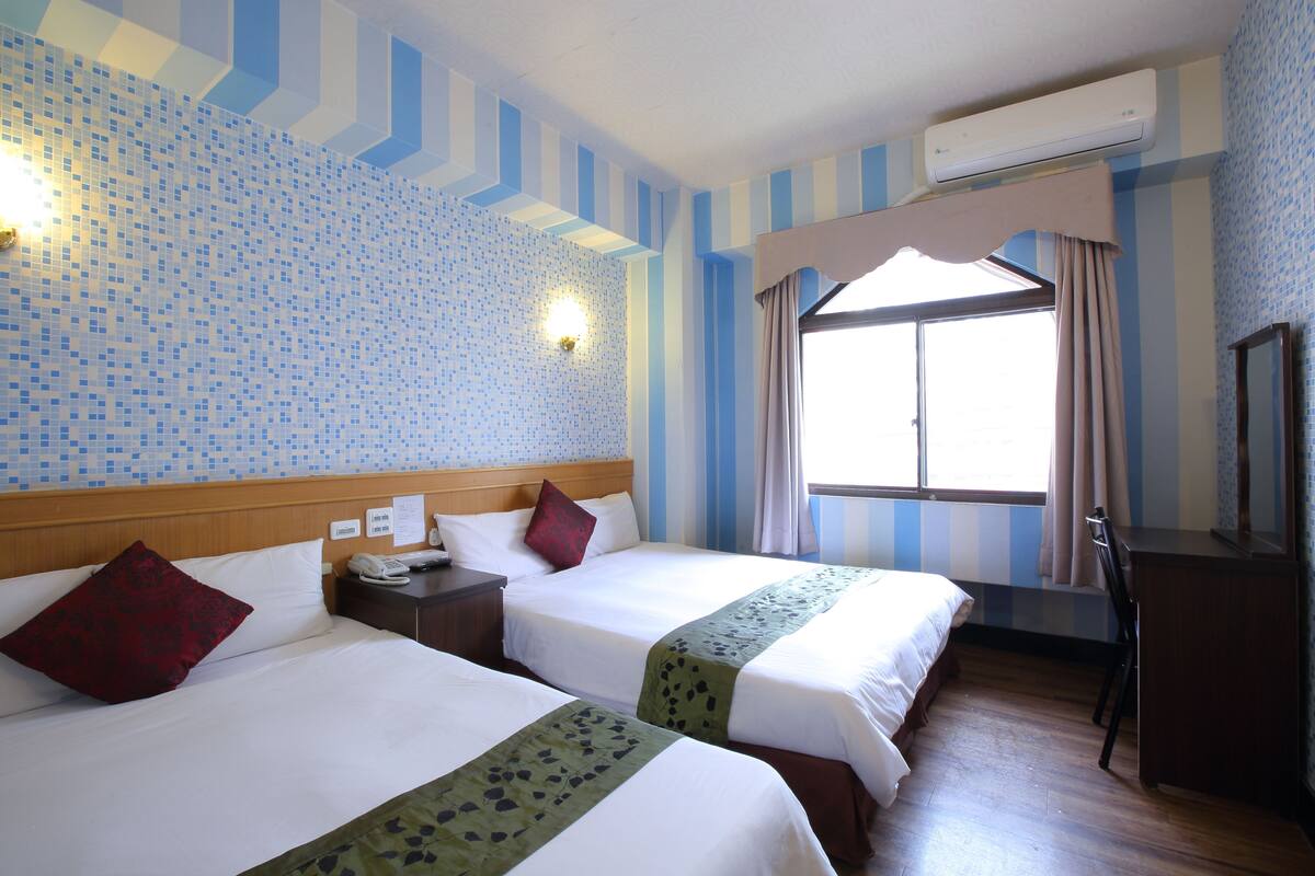 Habitación de hotel con dos camas, cabecera de madera, mesita de noche con teléfono, escritorio con silla y ventana con cortinas.