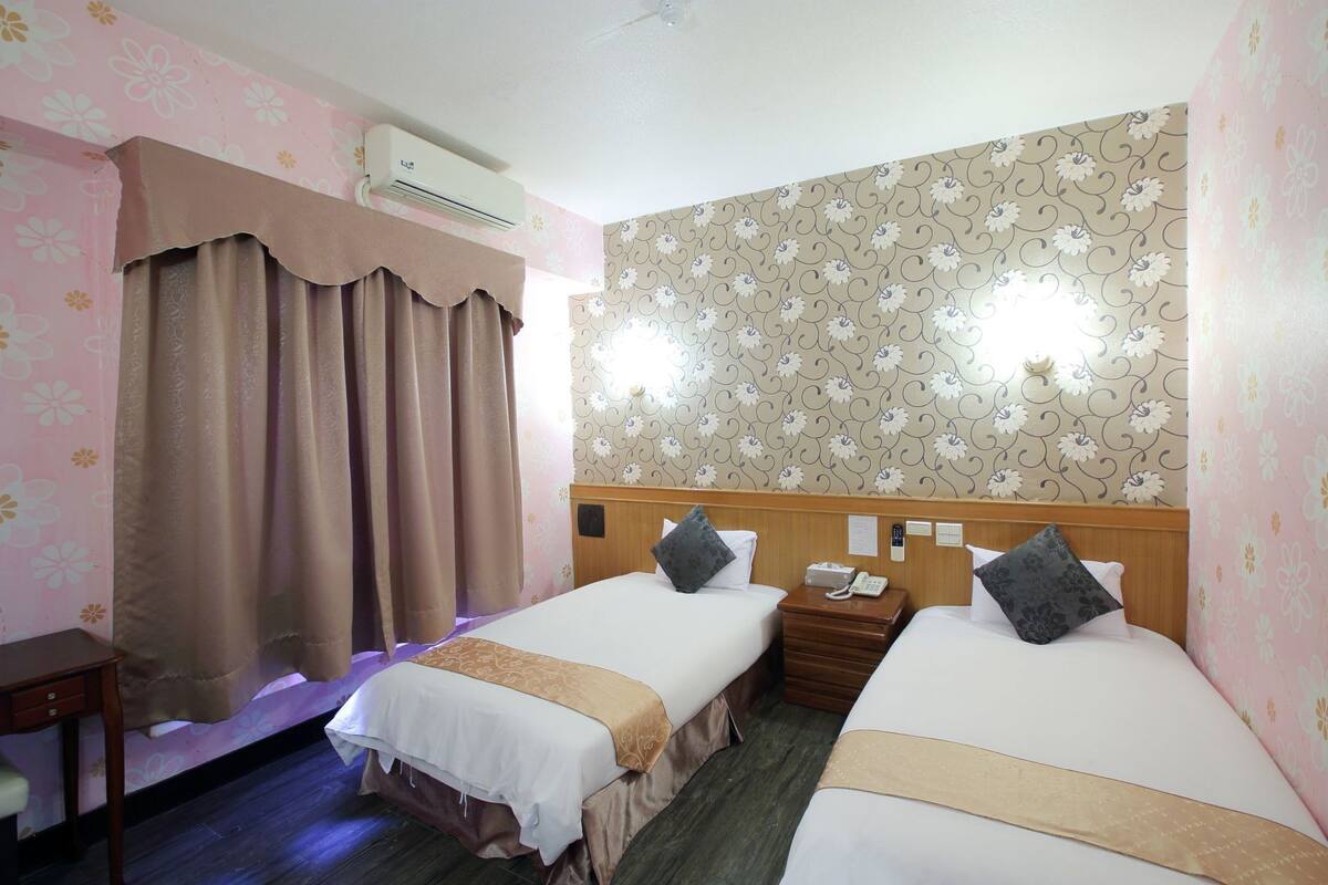 Habitación de hotel con dos camas, una mesita de noche con teléfono, aire acondicionado en la pared y una cortina.