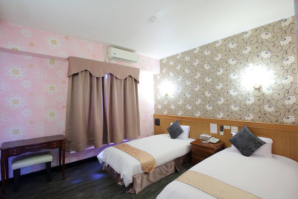 Habitación de hotel con dos camas, un escritorio y una silla. Hay un equipo de aire acondicionado sobre el escritorio y una lámpara de pared sobre las camas.