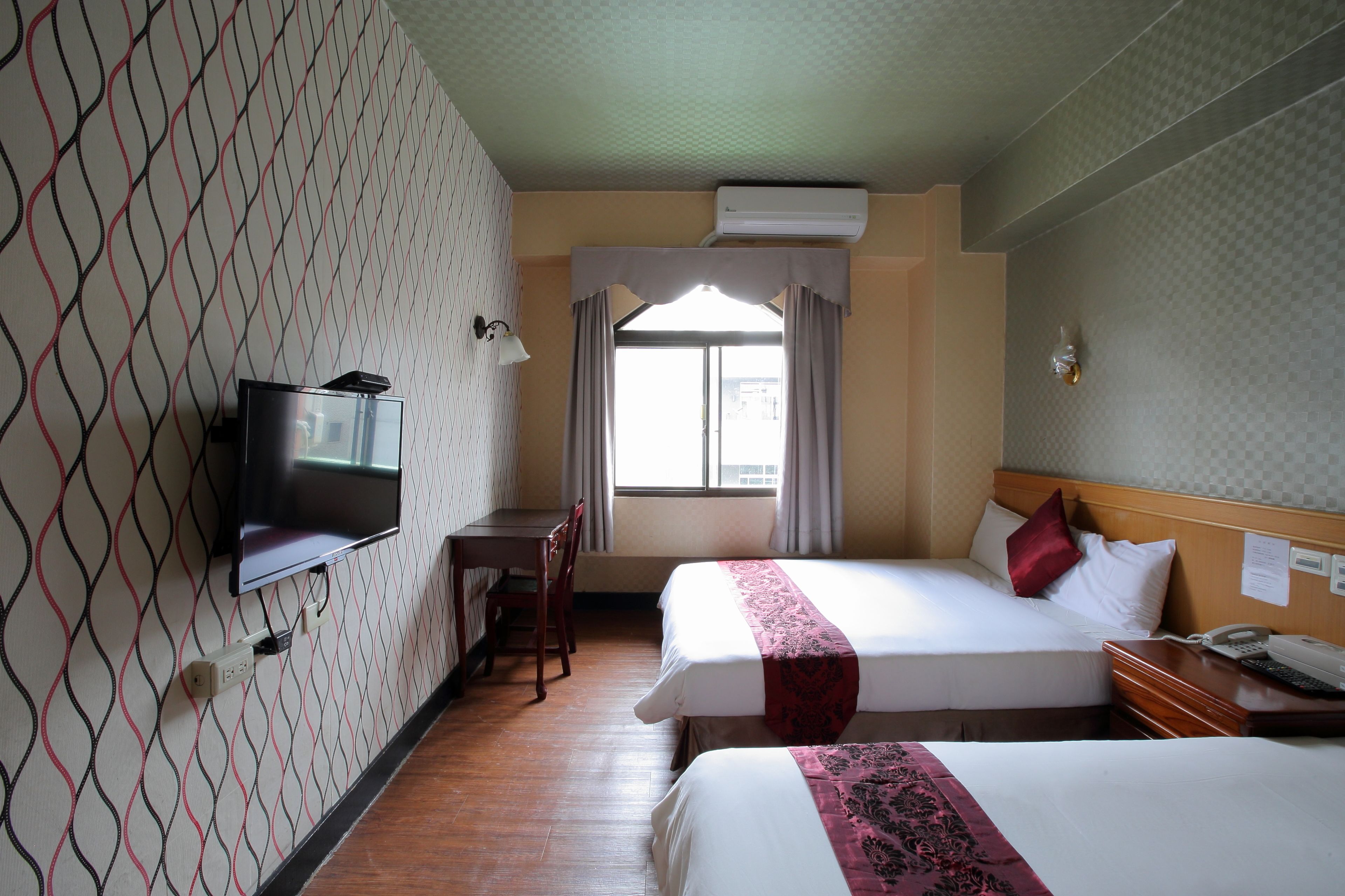A hotel room with two beds, a desk, a television, and a window with curtains.