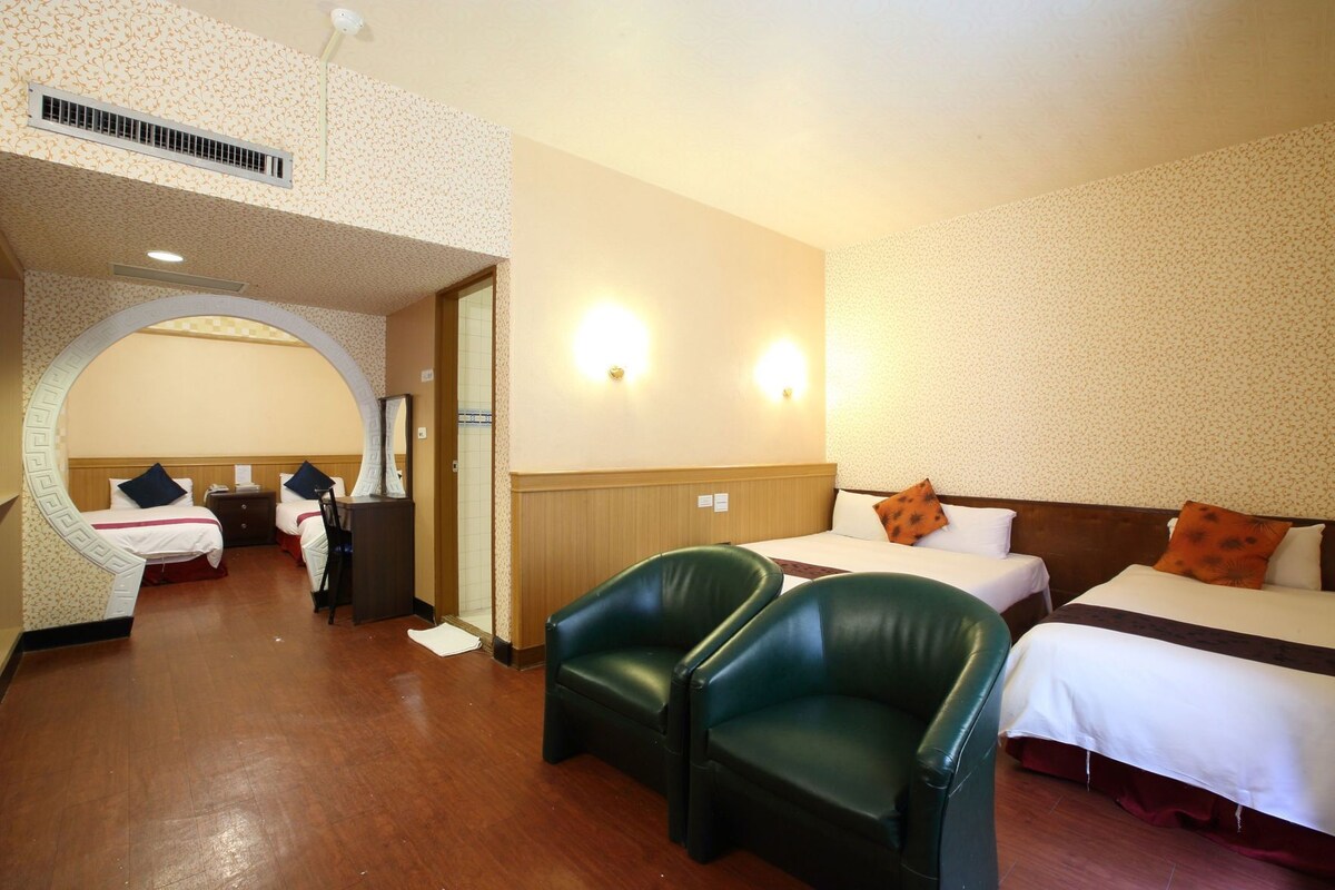 Una habitación de hotel con dos camas, un sillón de cuero verde, suelo de madera y pared con un estampado.
