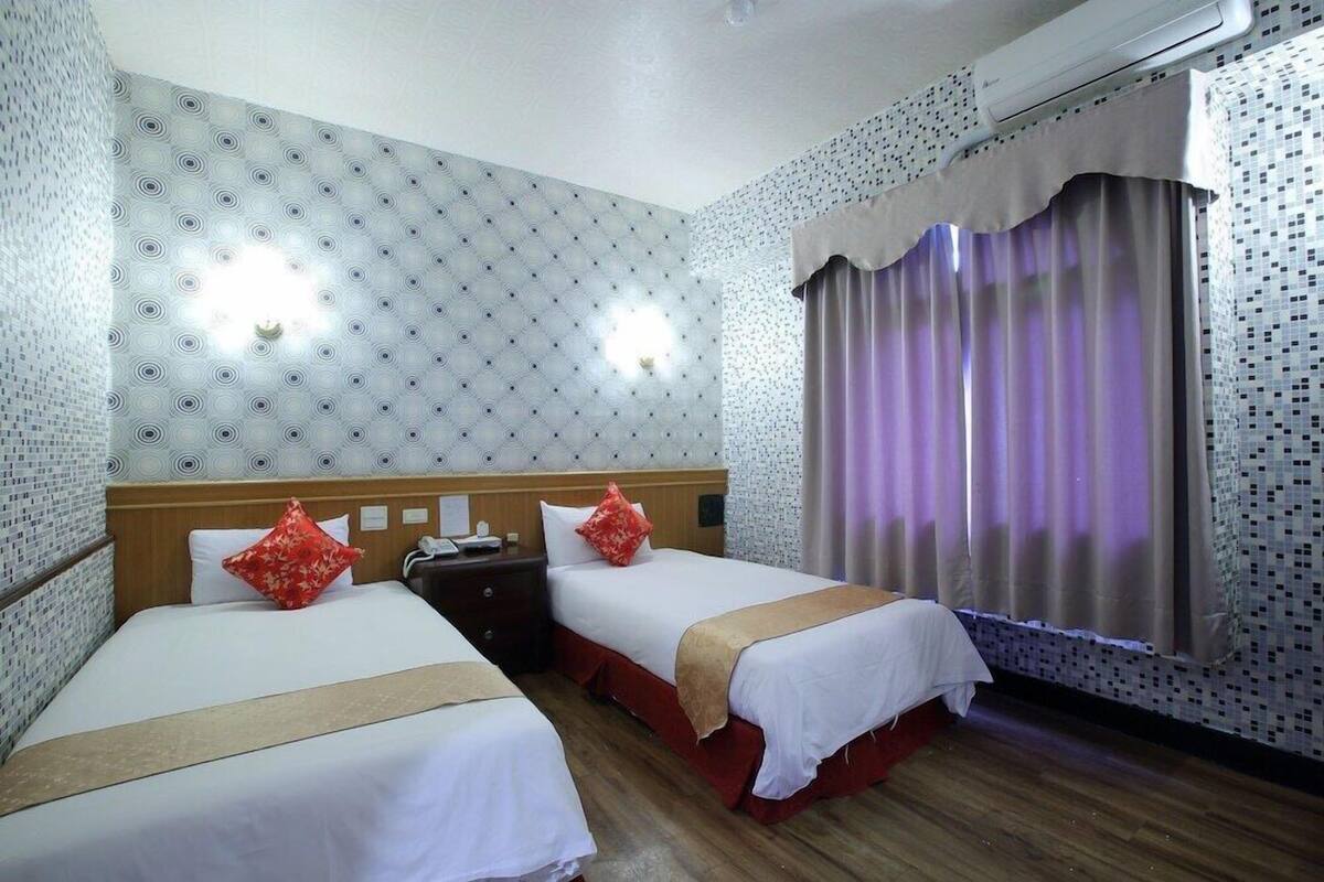 Habitación de hotel con dos camas, cabecero de madera, papel tapiz estampado, una ventana con cortinas y una mesita de noche.