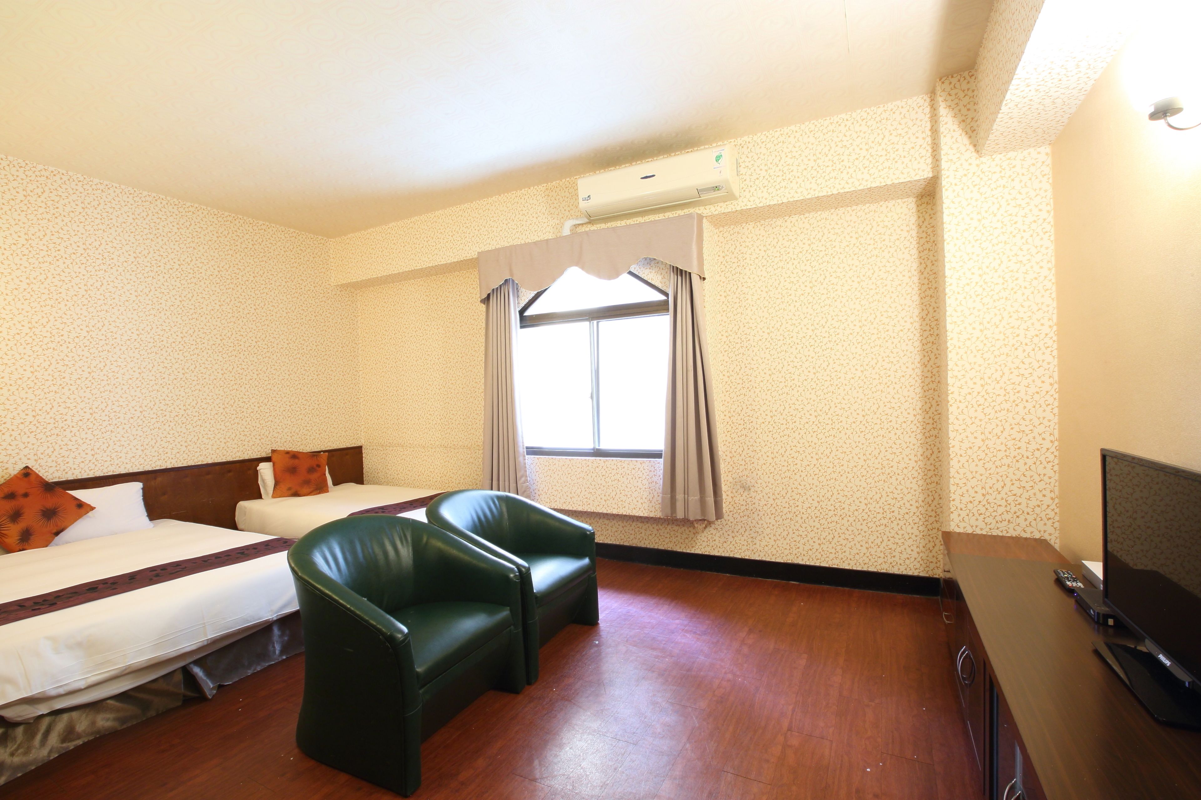 Ein Hotelzimmer mit zwei Betten, einem grünen Ledersessel, einem Holztisch mit Fernseher und einem Fenster mit Vorhängen.