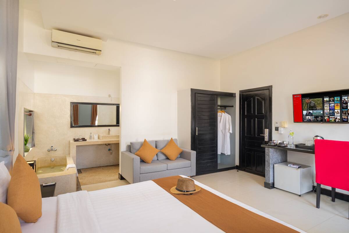 A hotel room with a bed, a sofa, a desk, a small fridge, a TV, and a bathroom with a sink and mirror.