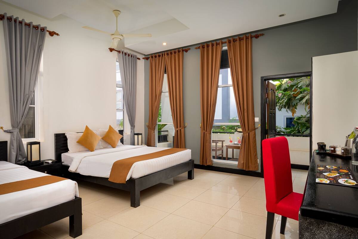 A hotel room with two beds, a red chair, a dining table with a menu, and a balcony with plants.