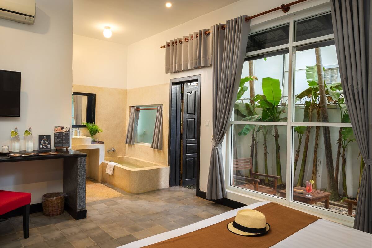 A modern bathroom with a bathtub, shower, and toilet. There is a sink with a mirror, a red chair, and a window with curtains.