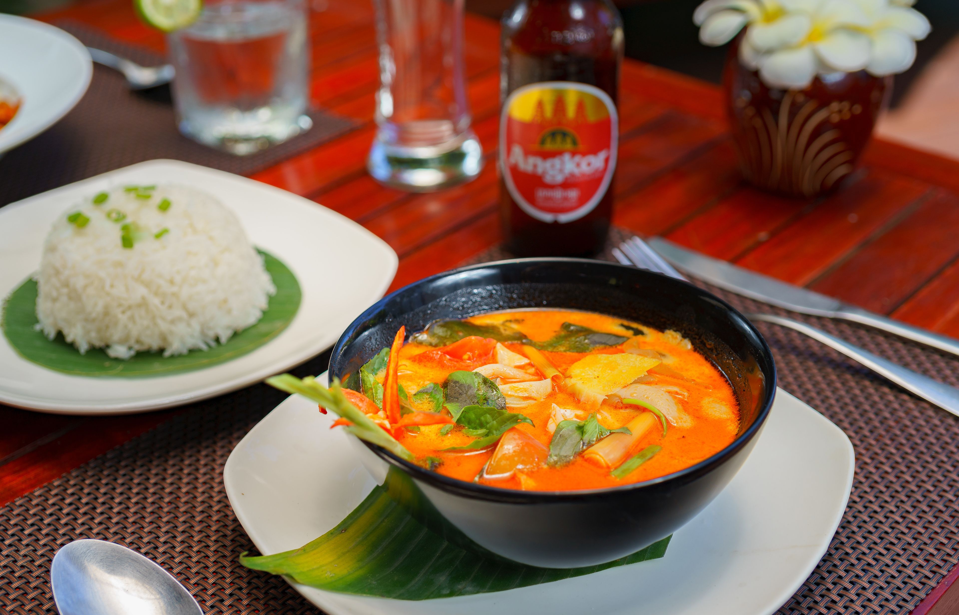 A table with a bowl of soup, a plate of rice, a bottle of sauce, and a glass of water.