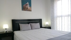 Una cama bien hecha con edredón blanco y dos almohadas, flanqueada por dos mesitas de noche con lámparas, en una habitación con un ventanal grande y un cuadro en la pared.