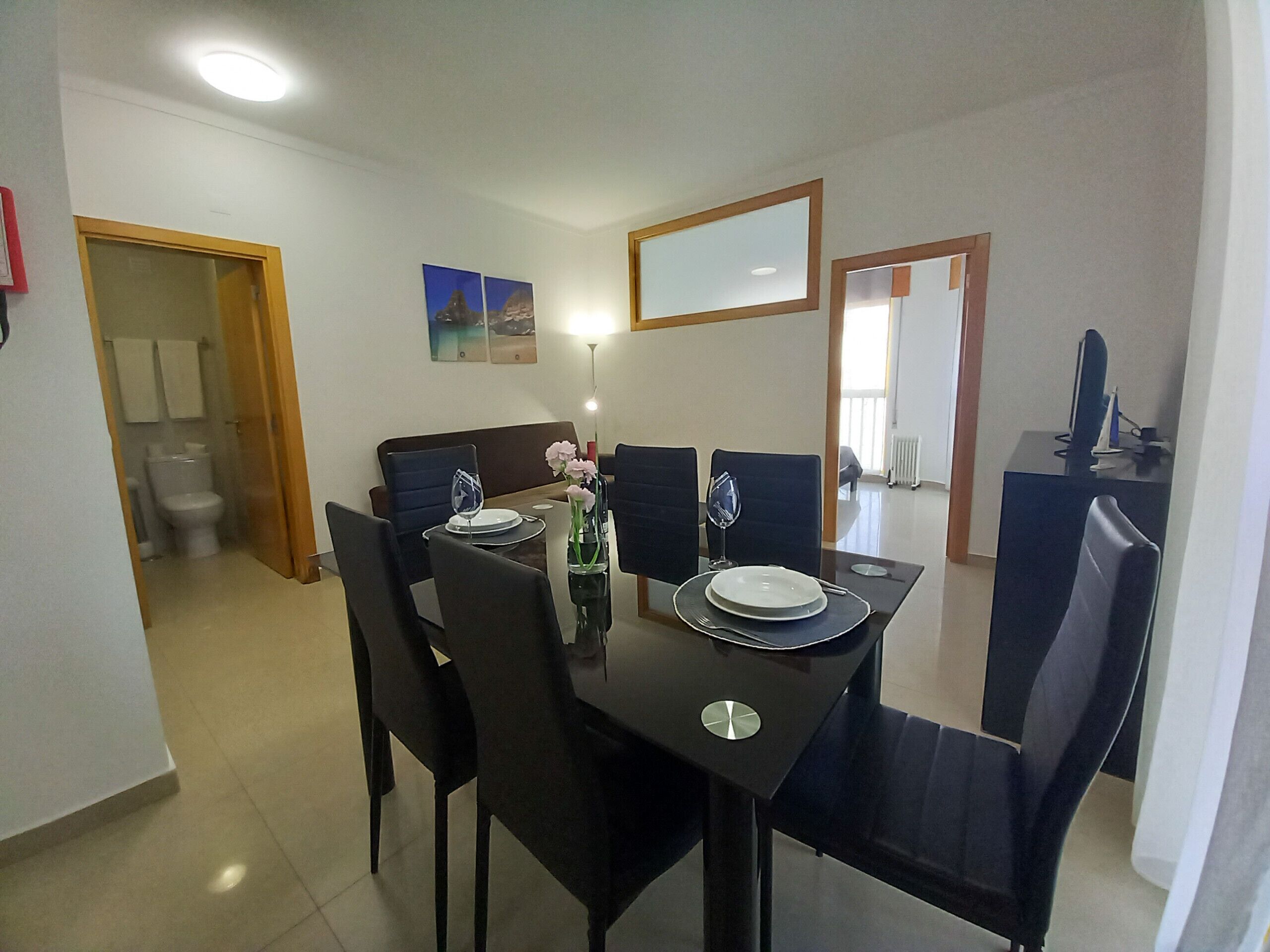 Un coin repas avec une table dressée pour quatre personnes, des chaises, un vase rempli de fleurs et un tableau accroché au mur.
