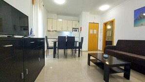 Una sala de estar moderna con un televisor de pantalla plana, un sofá oscuro y una mesa de centro. Se aprecia un comedor con mesa y sillas, y una cocina al fondo.