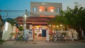 A two-story building with a white facade and a red-tiled roof. There are bicycles parked in front, and a white picket fence surrounds the entrance. - Cua Dai Beach Homestay (Da Nang)