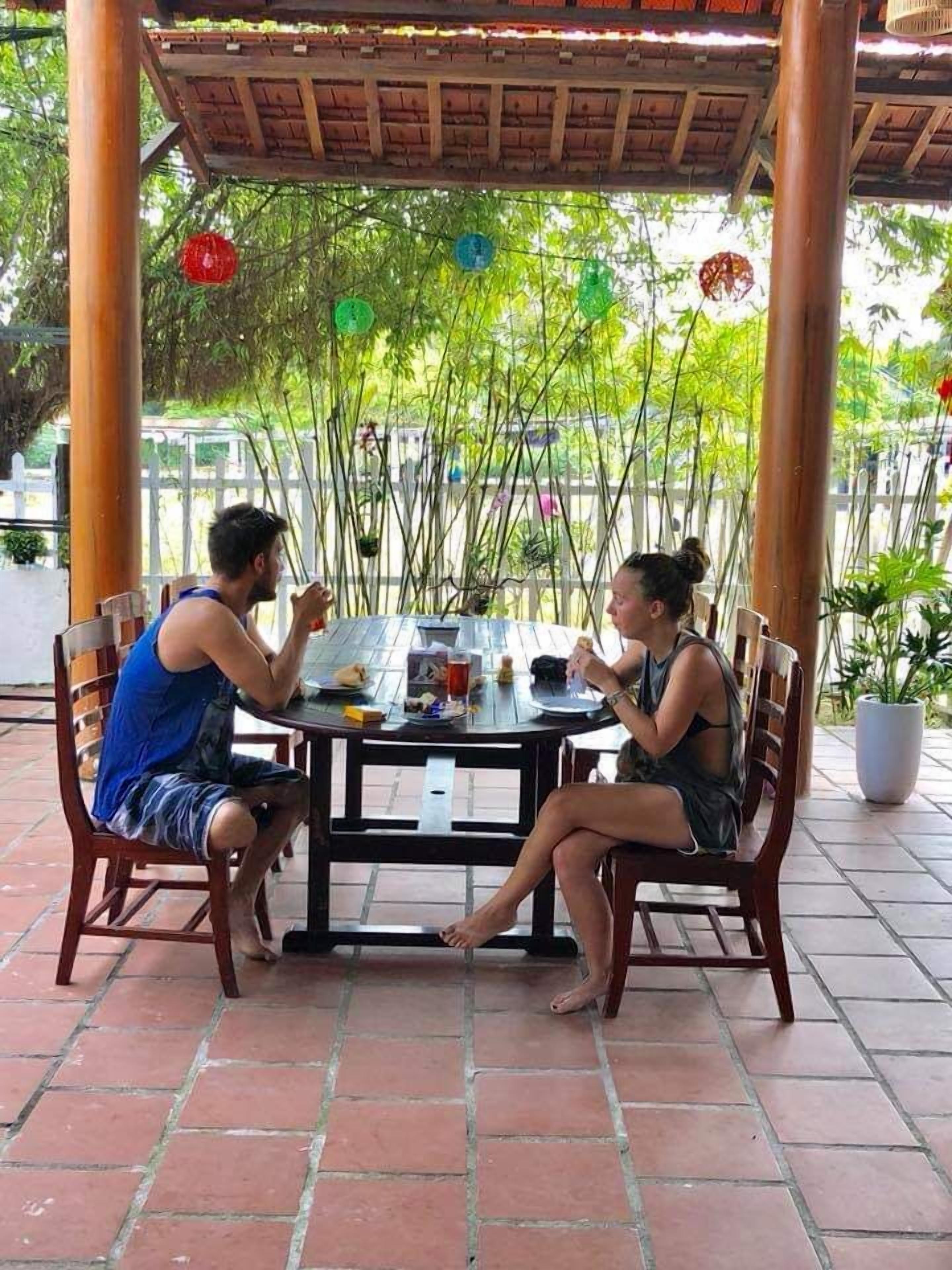 Duas pessoas sentadas à volta de uma mesa redonda sob uma pérgola de madeira, com soalho de azulejos, rodeadas por vegetação.