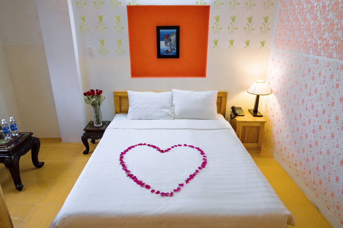 A hotel room with a neatly made bed, a heart-shaped flower arrangement on the bed, a bedside table with a vase of flowers, a nightstand with a lamp, and a picture on the wall.