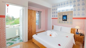 A hotel room with a bed, a nightstand, a wall-mounted TV, and a balcony with a view of greenery. - Cua Dai Beach Homestay (Da Nang)