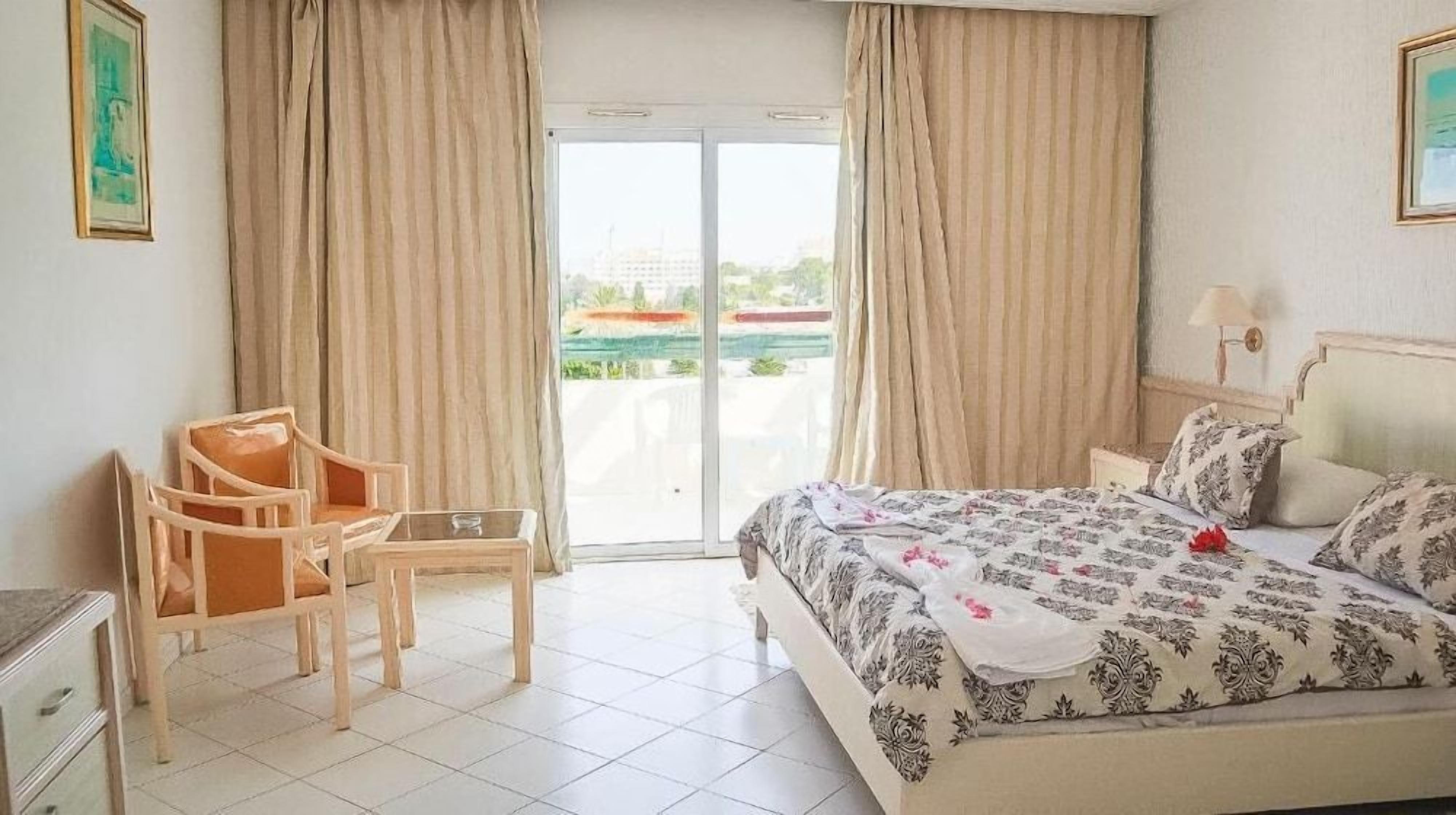 Habitació d'hotel amb un llit, una cadira, una taula petita i una vista a través d'una porta de vidre.