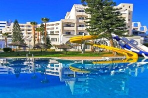 A swimming pool with a water slide, surrounded by a building with balconies and palm trees. - Hotel Royal Jinene (Sousse)