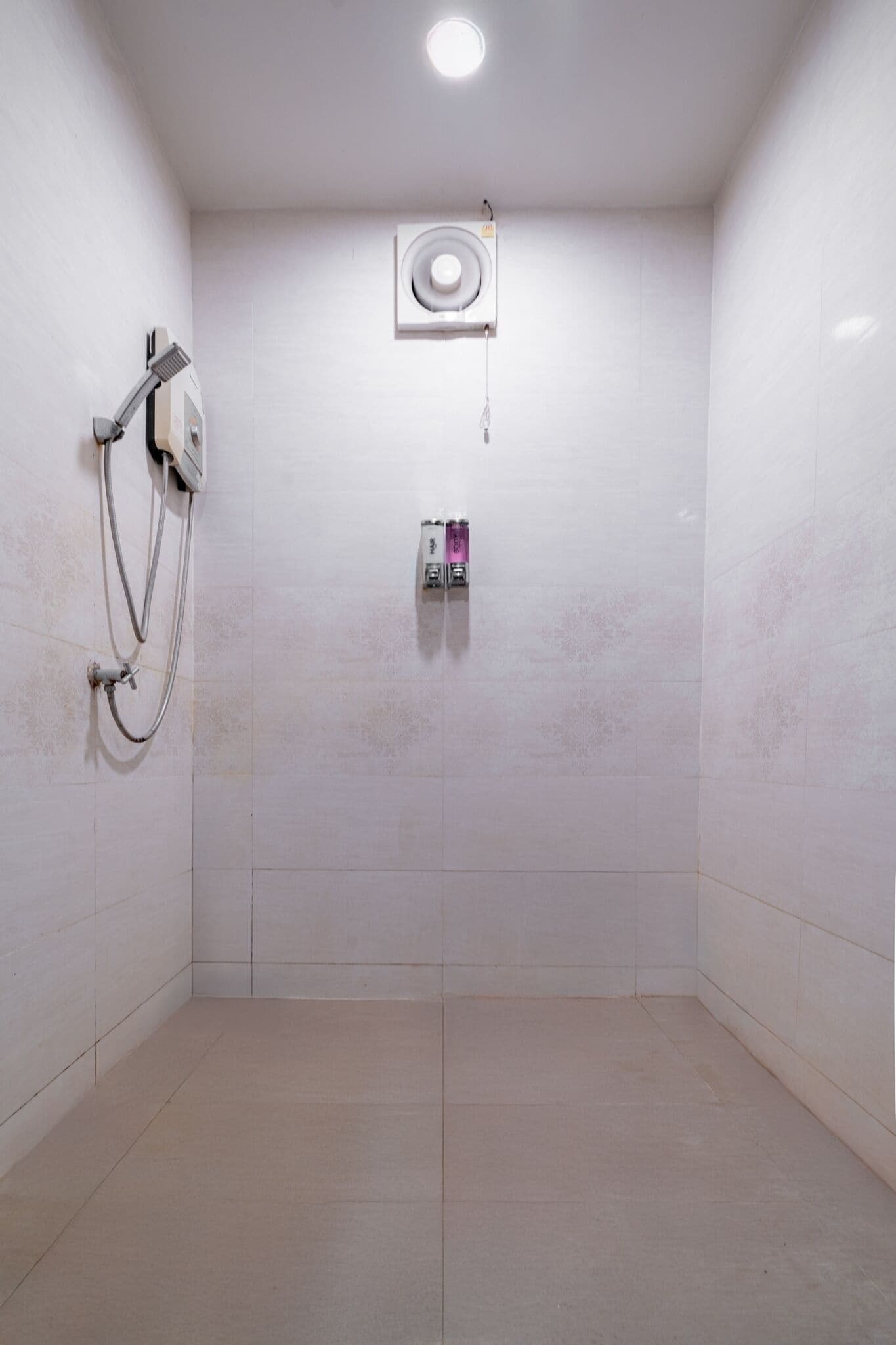 A small, tiled shower room with a handheld showerhead, a wall-mounted control panel, and a ceiling light.