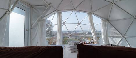 Habitación con estructura de techo geométrica, ventanas amplias, cama con ropa blanca y vistas al exterior.