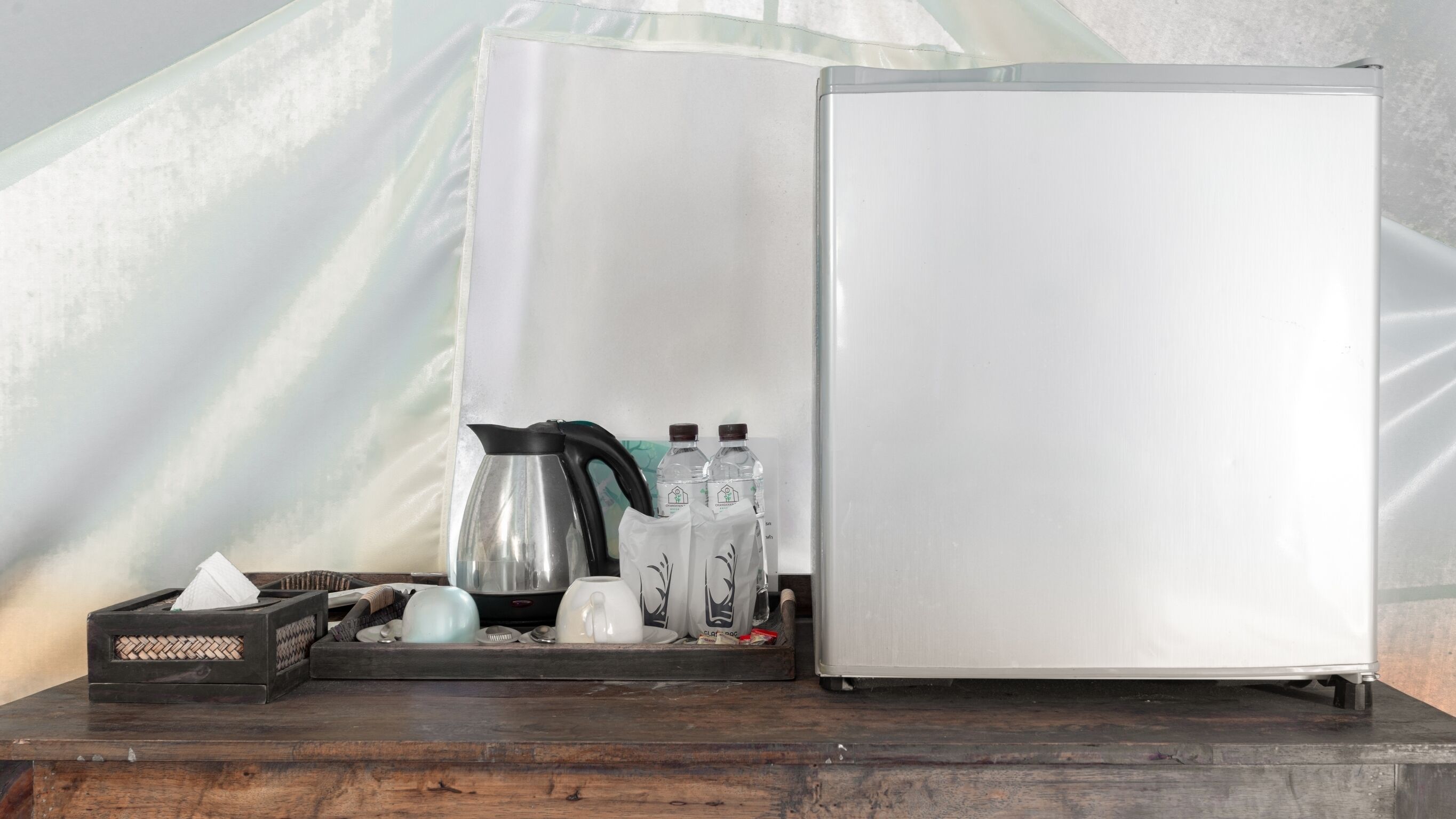 A compact refrigerator with a water dispenser, a kettle, and two bottles of water on a wooden surface.