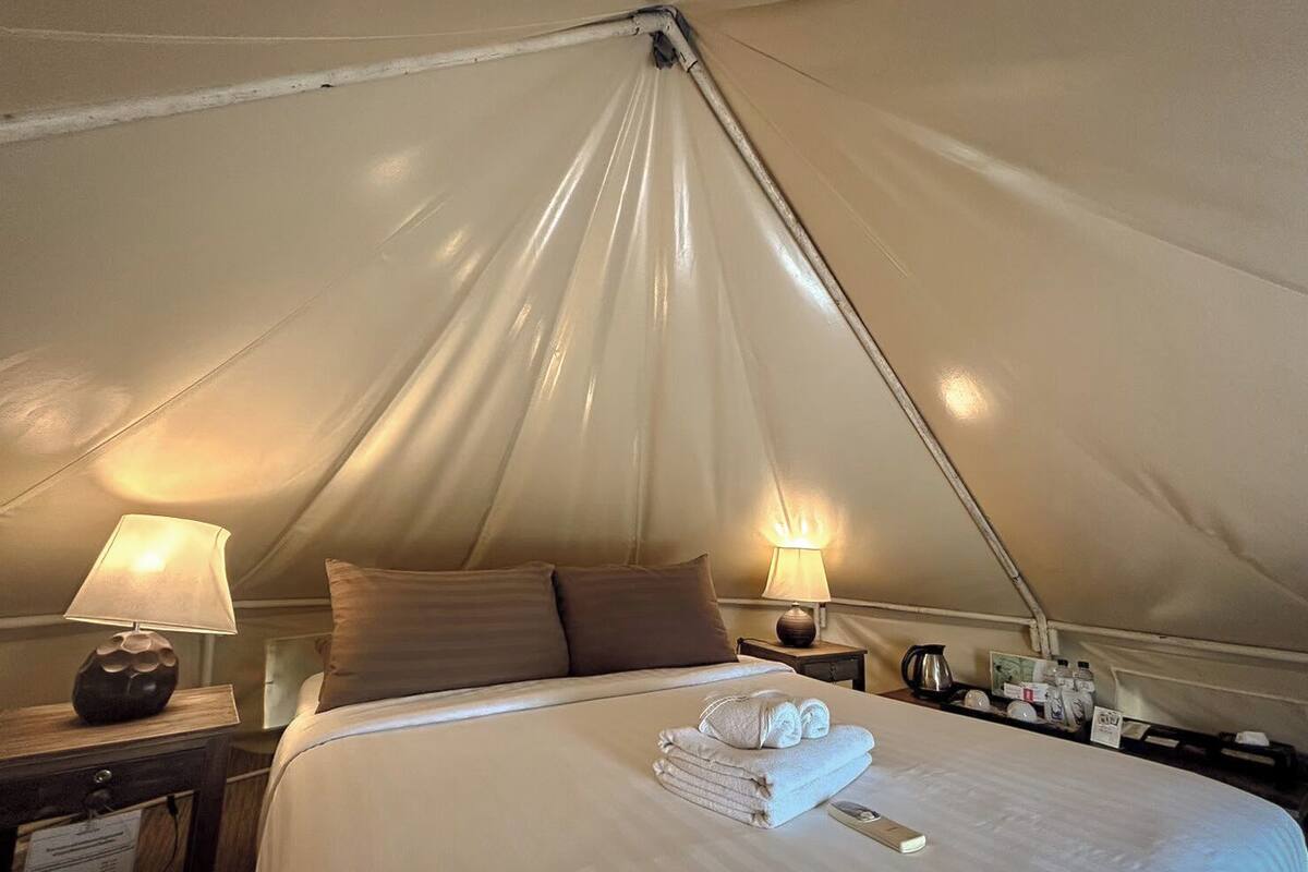 A tented room with a bed, two bedside lamps, a nightstand with a lamp, and a folded towel on the bed.