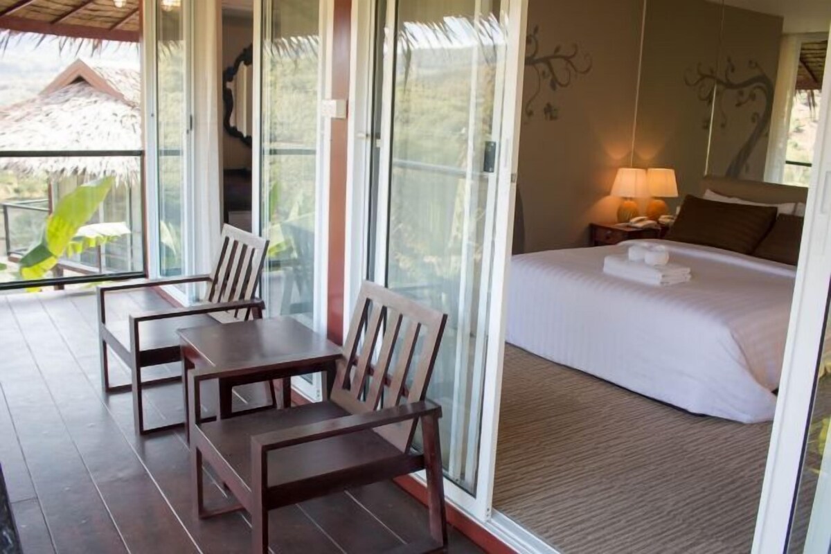 A room with a bed, a nightstand with a lamp, a table with chairs, and a sliding glass door leading to a balcony.