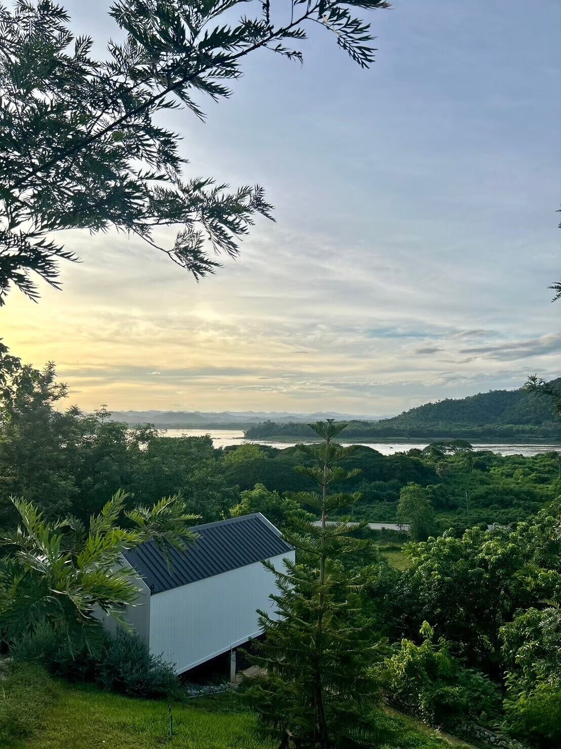 A modern house with a sloped roof, surrounded by lush greenery and a view of a lake and distant mountains.