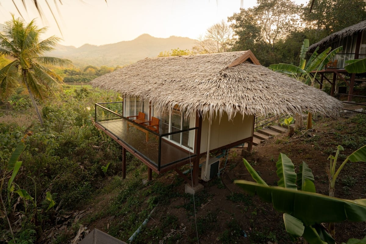 A thatched-roof structure with a glass balcony, surrounded by lush greenery and mountains in the background.