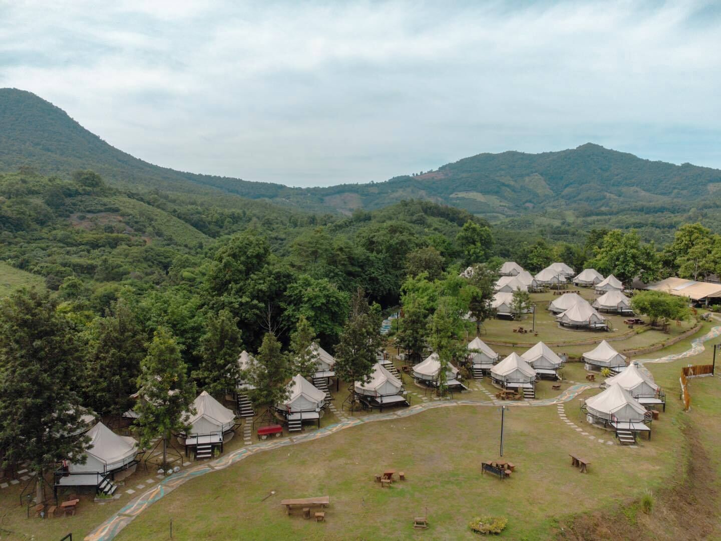 A campsite with multiple tents arranged in rows, surrounded by lush greenery and hills in the background.