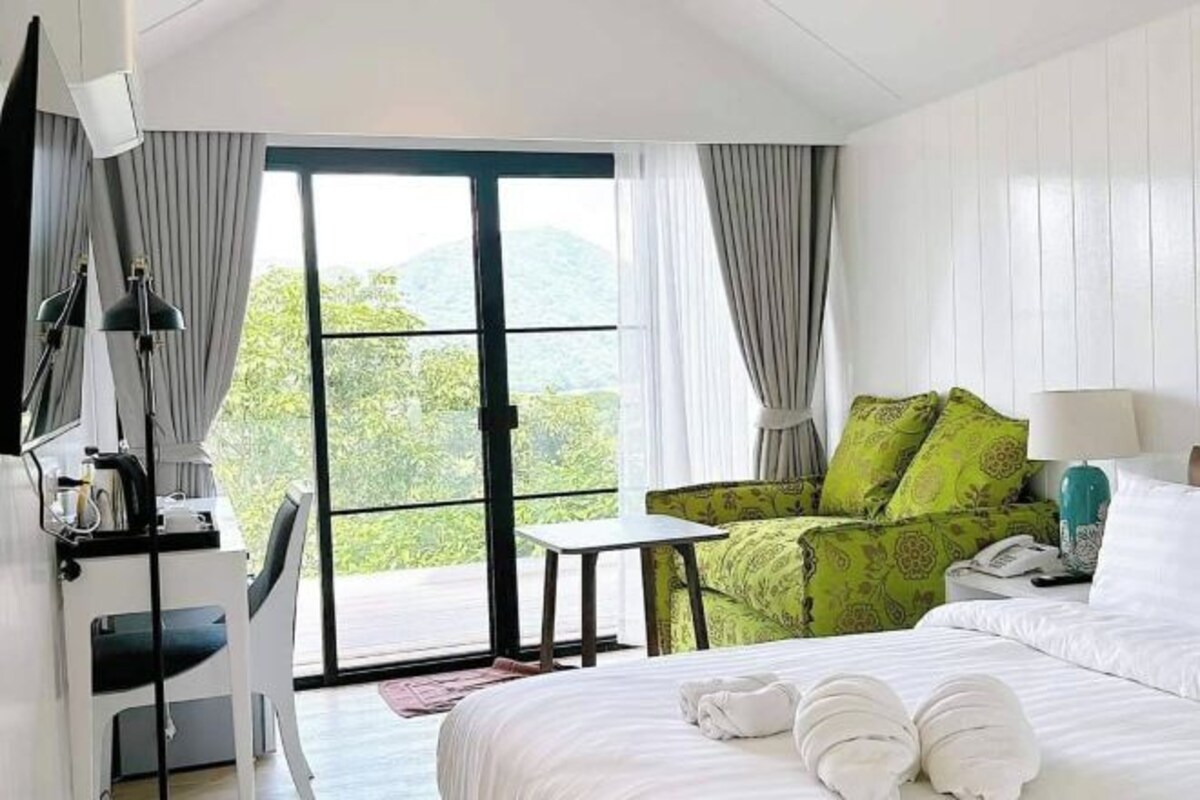 A hotel room with a bed, a desk, a chair, and a view of greenery through the window.