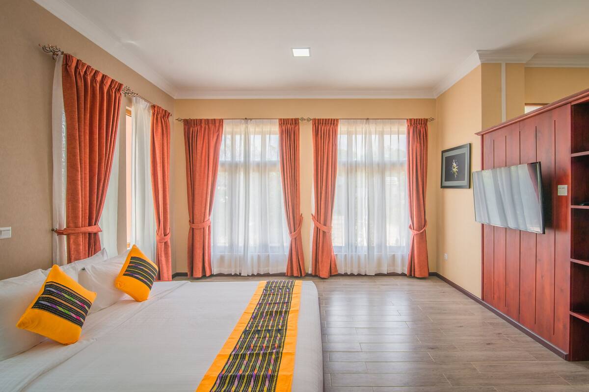 A hotel room with a bed, orange curtains, a TV, and a wooden wardrobe.