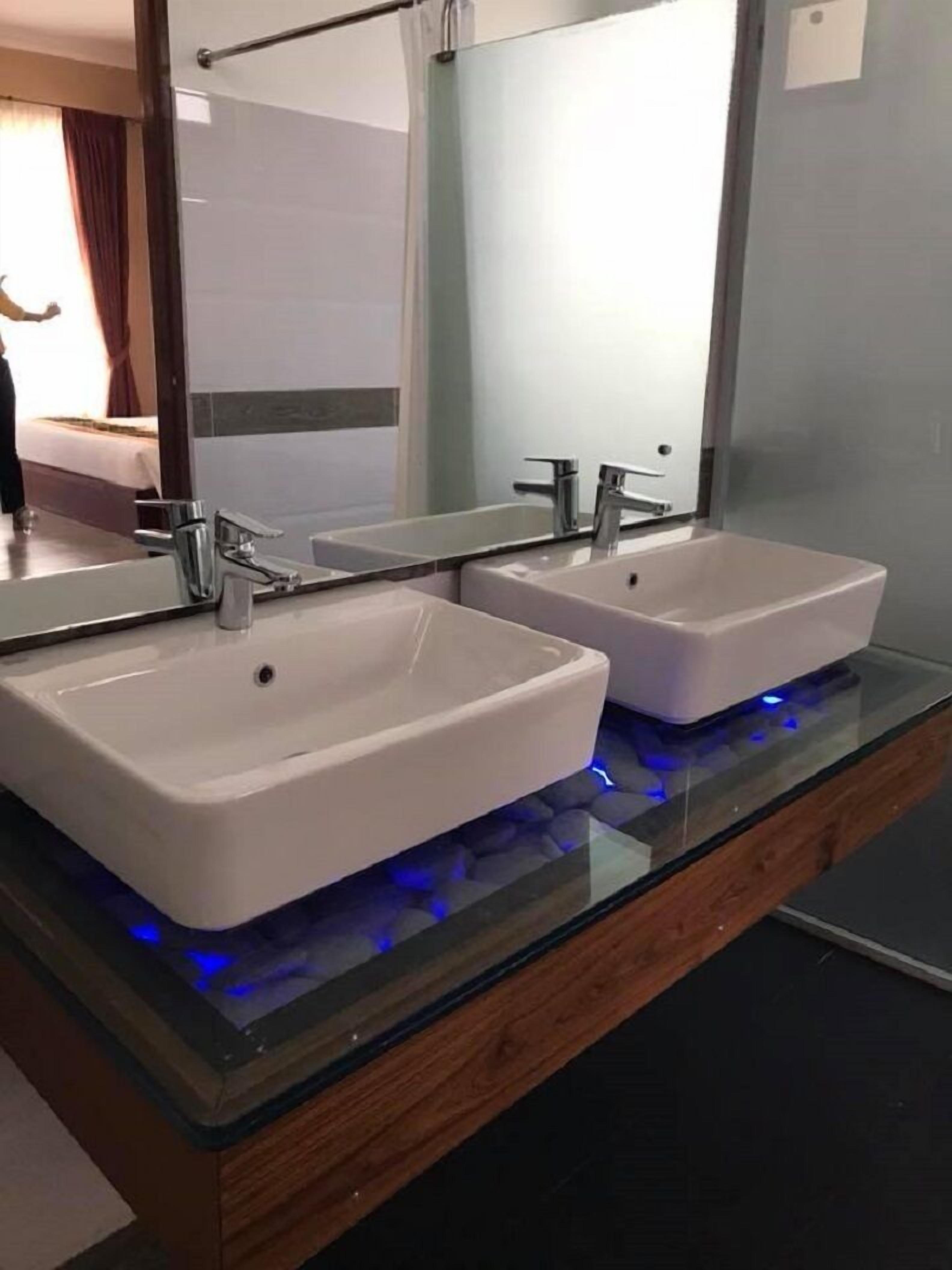 Un baño moderno con dos lavamanos blancos, un espejo grande y una encimera de vidrio con iluminación azul.