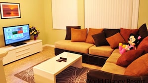 Una sala de estar con un sofá color beige, una mesa de centro, un televisor sobre un mueble blanco y un peluche de Mickey Mouse.