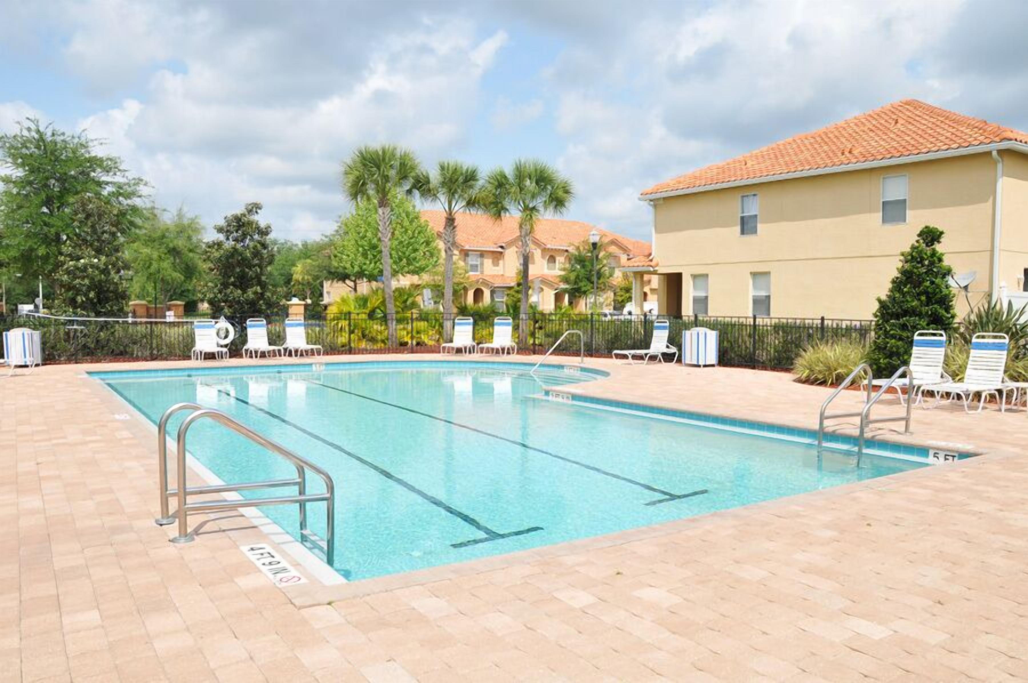 A swimming pool with a tiled deck, lounge chairs, and a building with a tiled roof in the background.